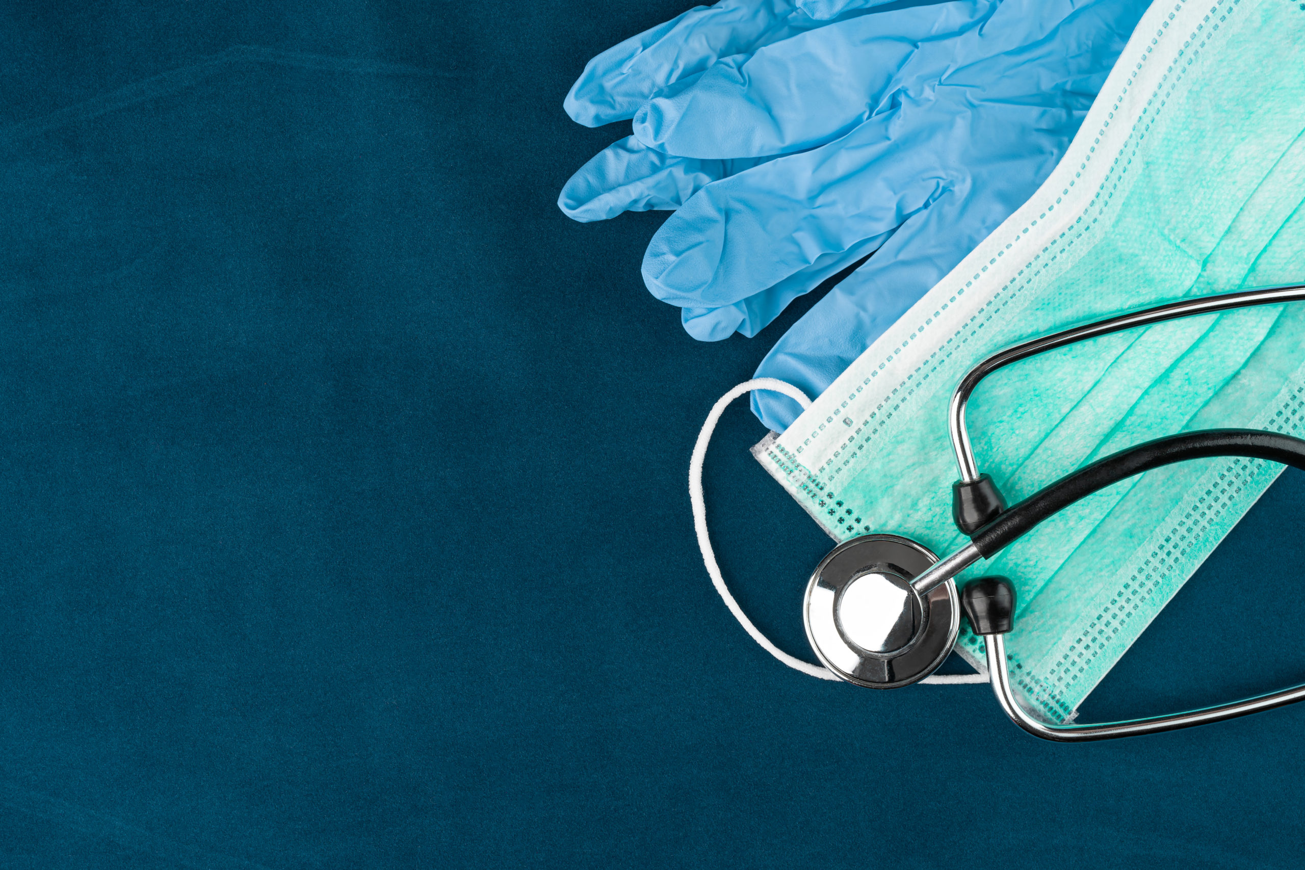 Green protective face mask, stethoscope and gloves on a blue background. Copy space on the left.