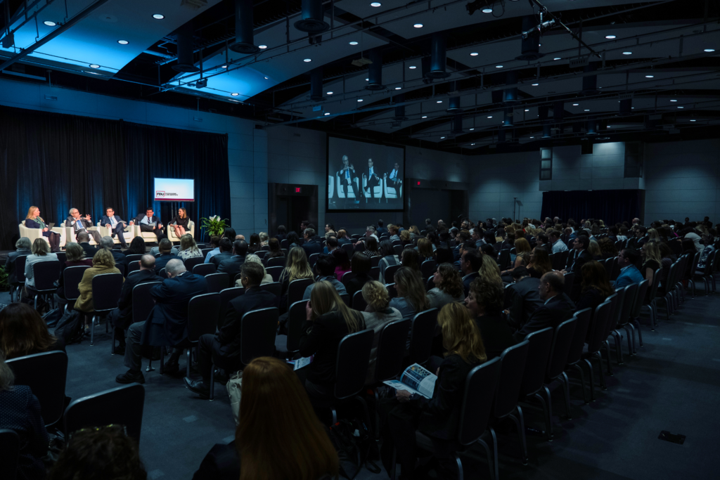 Image of the annual conference - an auditorium of people staring at a lit stage with 5 panelists.