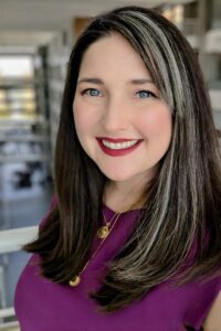 Pictured: A smiling woman with light beige skin, blue eyes, and dark brown hair, wears a purple top.