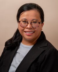Pictured: A smiling woman with light sienna skin, black hair pulled back in a ponytail, tortoiseshell glasses, and a pink lip-tint, wearing a black coat and grey top.