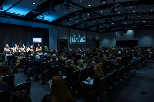 Image of the annual conference - an auditorium of people staring at a lit stage with 5 panelists.