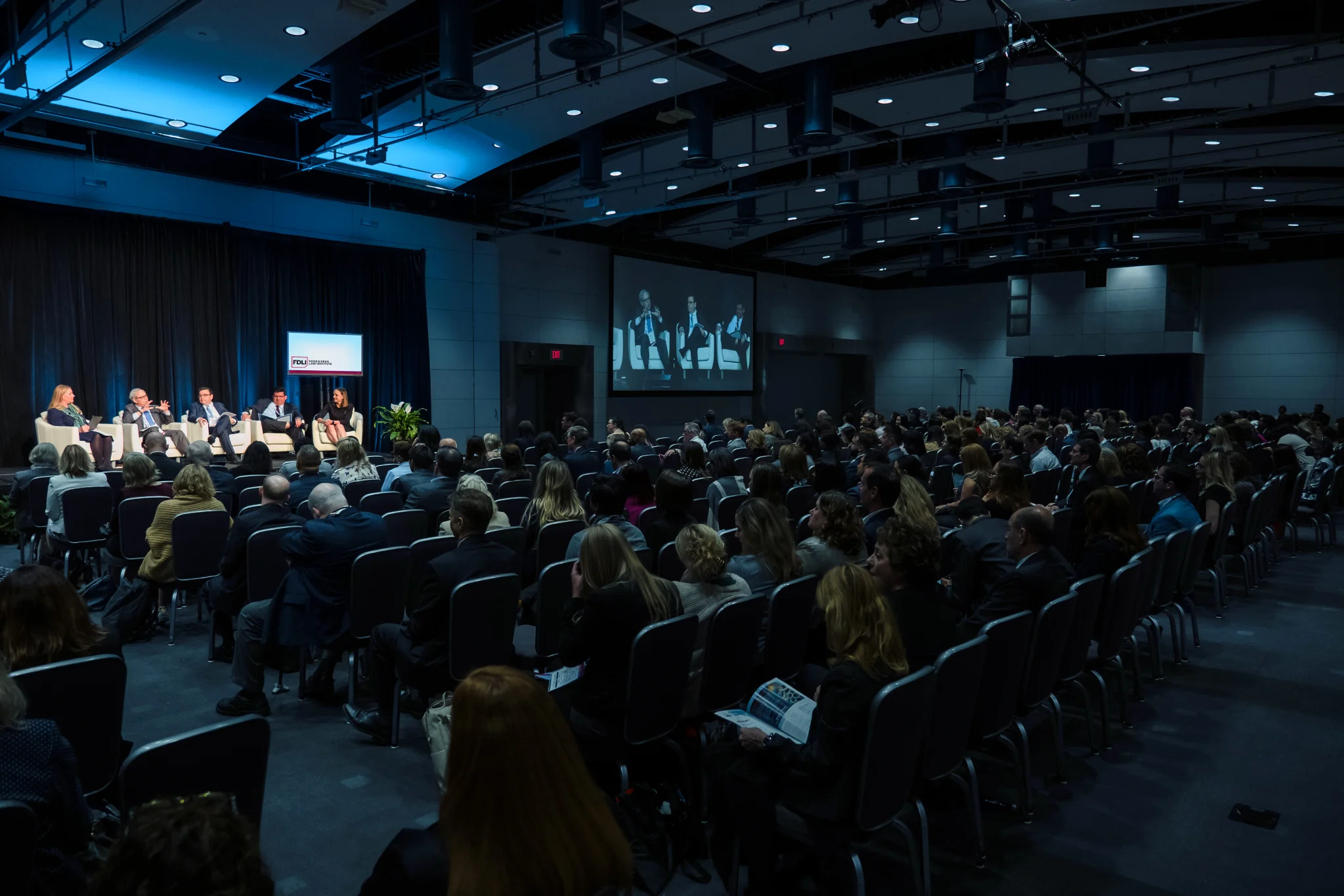 Image of the annual conference - an auditorium of people staring at a lit stage with 5 panelists.